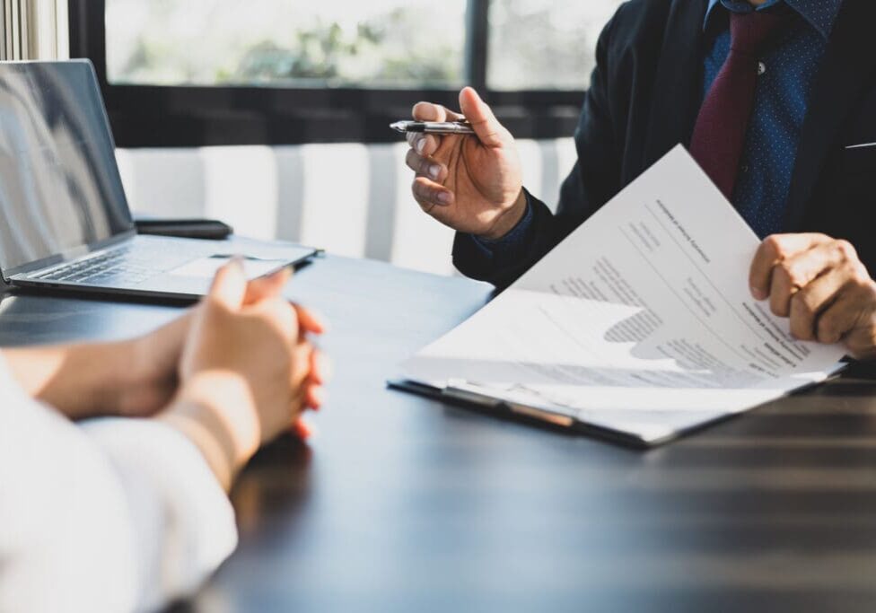 Businessman in suit in his office showing an insurance policy and pointing with a pen where the policyholder must to sign. Insurance agent presentation and consulting insurance detail to customer.